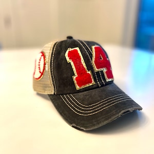 May include: A charcoal gray baseball cap featuring a distressed finish and a curved brim with white stitching. The front displays the number 14 in red fuzzy fabric with gold trim, while the side panel features a white baseball graphic.