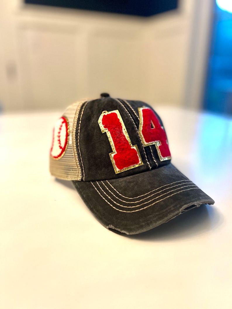 May include: A charcoal gray baseball cap featuring a distressed finish and a curved brim with white stitching. The front displays the number 14 in red fuzzy fabric with gold trim, while the side panel features a white baseball graphic.