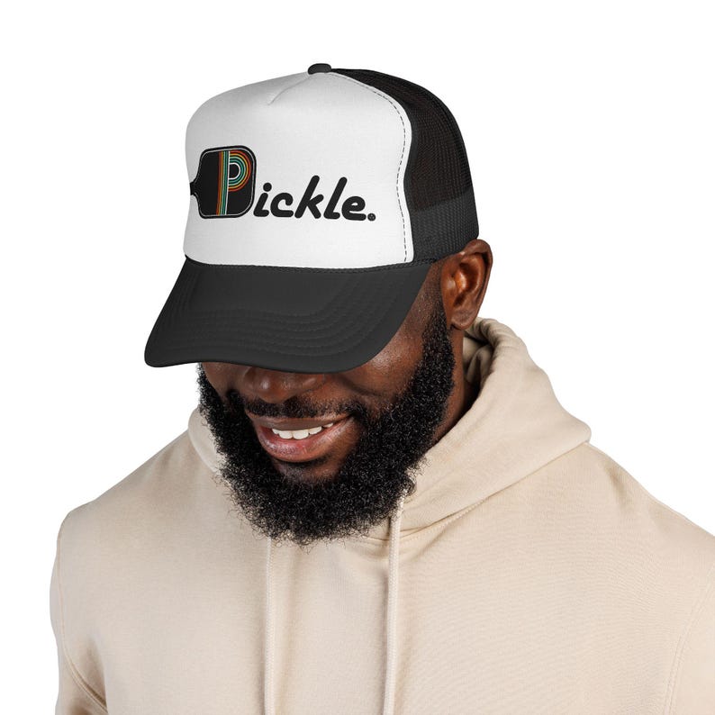 May include: A black and white trucker hat featuring a graphic of a pickleball paddle shaped like the letter P, followed by the text "ickle." in black. The hat has a white front panel, a black brim, and a black mesh back for ventilation.