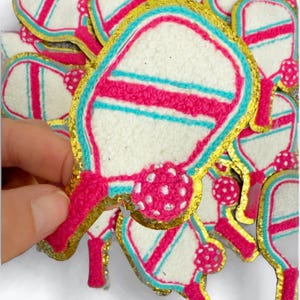 May include: A stack of pink and blue paddleball rackets with gold glitter trim. The rackets are made of white felt and have a pink and blue striped design. Each racket has a pink ball with white dots.