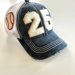 May include: A blue baseball cap with a white mesh back and a baseball patch. The cap features the number 25 in white with gold trim. The brim has white stitching.