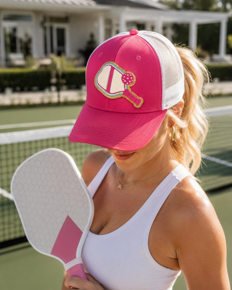 Peut inclure: Une casquette de type trucker rose vif avec un filet blanc &agrave; l'arri&egrave;re et un &eacute;cusson central repr&eacute;sentant une raquette et une balle de pickleball. L'&eacute;cusson pr&eacute;sente une bordure dor&eacute;e scintillante, une raquette cr&egrave;me et une balle rose &agrave; pois.