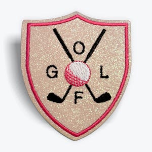 May include: A glittery beige shield-shaped patch with a pink border. The patch features the word "GOLF" with a golf ball and crossed golf clubs in black. The golf ball is white and pink.