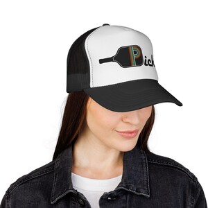 May include: A black and white trucker hat featuring a graphic of a black pickleball paddle with a rainbow-striped letter P and the text "ick" in black lettering. The hat has a solid white front panel, a black brim, and black mesh back panels.