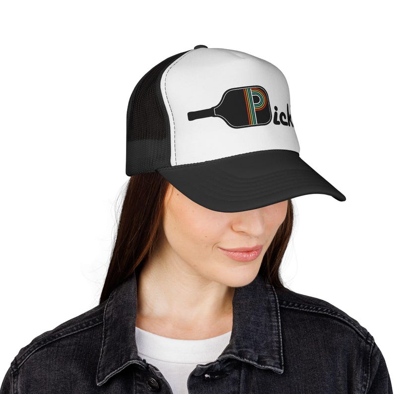 May include: A black and white trucker hat featuring a graphic of a black pickleball paddle with a rainbow-striped letter P and the text "ick" in black lettering. The hat has a solid white front panel, a black brim, and black mesh back panels.