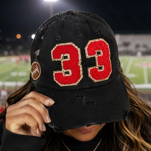 May include: A black distressed trucker hat featuring the number 33 in red felt with gold glitter trim on the front panels. A small brown football-shaped patch is stitched onto the side of the cap, which has a mesh back and a curved brim.