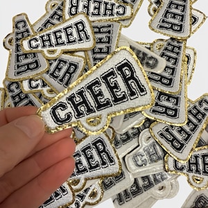 May include: A pile of white and gold iron-on patches with the word "CHEER" in black lettering. The patches are shaped like megaphones.