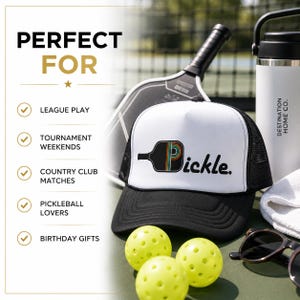 May include: A white and black trucker hat featuring a graphic of a pickleball paddle with a rainbow-striped center and the word pickle in black text. The hat is displayed with three yellow pickleballs, a paddle, a white water bottle, and sunglasses.