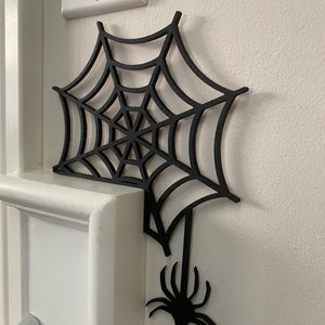 May include: Black spider web cutout with a spider hanging from the bottom. The web is attached to a white wall.