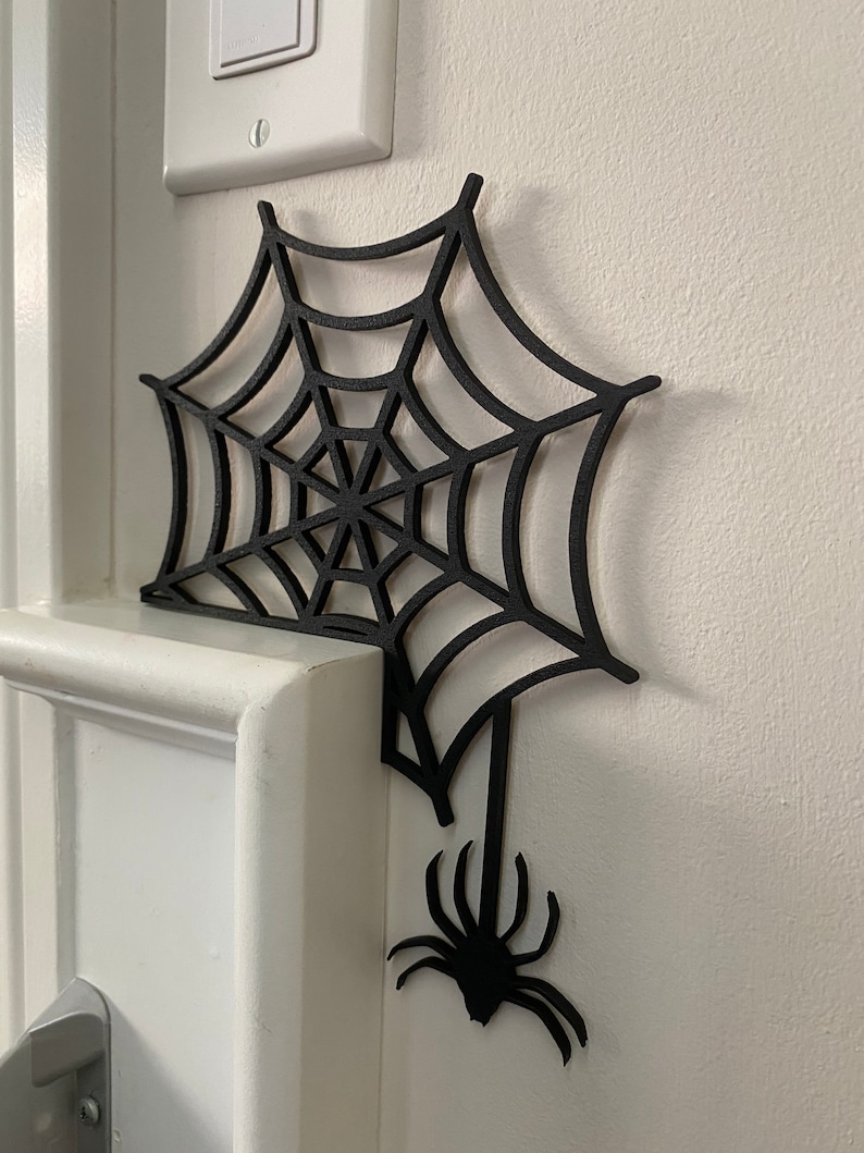 May include: Black spider web cutout with a spider hanging from the bottom. The web is attached to a white wall.