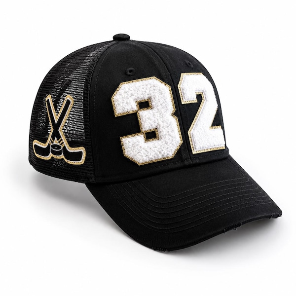 Puede incluir: Una gorra de béisbol negra con parte trasera de malla y visera curva. El frente muestra el número 32 en letras grandes, blancas y texturizadas de estilo chenilla con borde dorado. Un gráfico de dos palos de hockey cruzados está en el lateral.