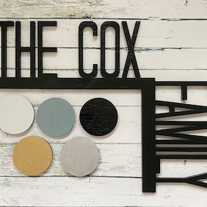 May include: Black wooden sign with the text "The Cox Family" in large, bold letters. The sign is decorated with six round, colorful coasters.
