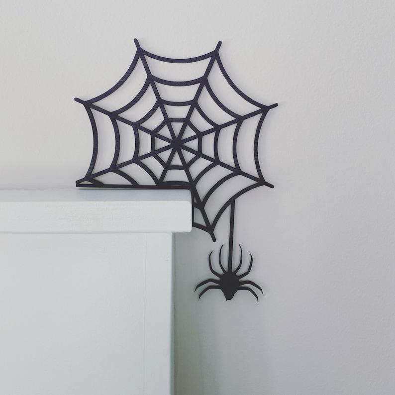 May include: Black spider web and spider cutout decoration for Halloween. The web is attached to a white shelf.