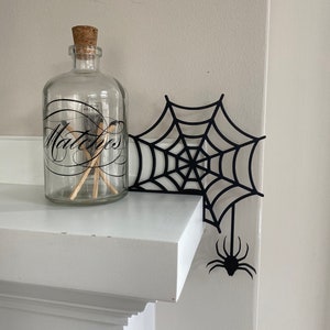 May include: A black spider web cutout with a spider hanging from the bottom. The web is placed on a white mantel with a glass jar labeled "Matches" on the left side.