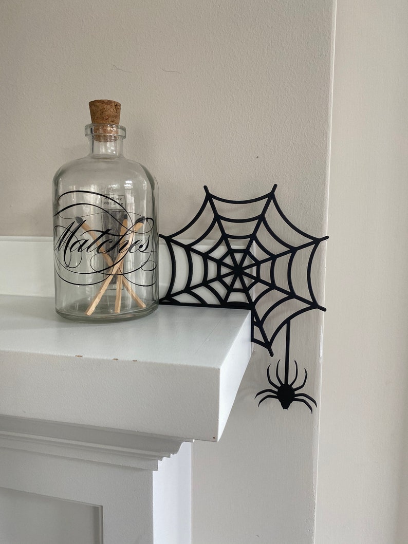 May include: A black spider web cutout with a spider hanging from the bottom. The web is placed on a white mantel with a glass jar labeled "Matches" on the left side.