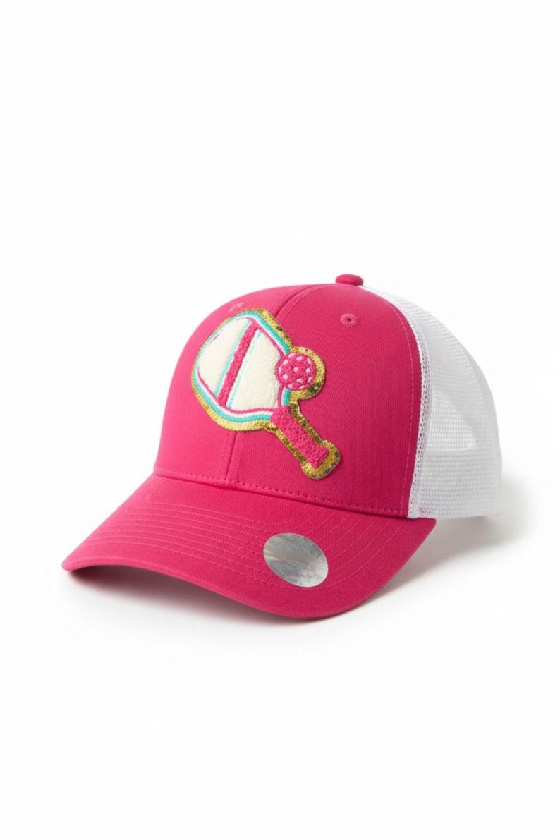 Peut inclure: Une casquette de type trucker rose vif avec un filet arri&egrave;re blanc et un &eacute;cusson frontal repr&eacute;sentant une raquette et une balle de pickleball. La raquette est bord&eacute;e d'or avec un centre blanc, la balle est rose &agrave; pois blancs. Un autocollant argent&eacute; est sur la visi&egrave;re.