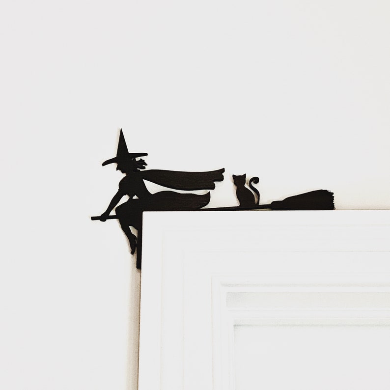 May include: Black silhouette of a witch flying on a broomstick with a black cat sitting on the broom handle. The silhouette is mounted on a white door frame.