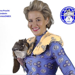 May include: A woman with blonde hair is smiling and holding a stuffed sugar glider. She is wearing a blue shirt with a yellow neck warmer and a pattern of blue and white circles. The shirt has a logo on it that says "PAD AURISTA.COM" and "MED".