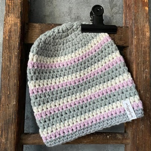 Puede incluir: Gorro de punto de crochet a rayas grises, blancas y rosas con una pequeña etiqueta blanca que dice "foggydew".