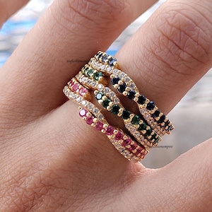 May include: A collection of gold-toned rings, each adorned with a row of colorful gemstones. The rings feature ruby, emerald, sapphire, and clear stones. The rings are stacked on a finger, showcasing the jewelry's design.
