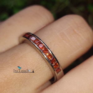 Puede incluir: Un anillo de banda de plata con una fila de piedras preciosas naranjas. El anillo está en un dedo.