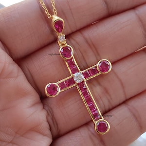 Op de afbeelding: Een goudkleurige hangerketting met een kruisontwerp. Het kruis is versierd met heldere rode edelstenen en een centrale heldere steen. De ketting heeft een fijne ketting en een druppelvormige edelsteen aan de bovenkant.