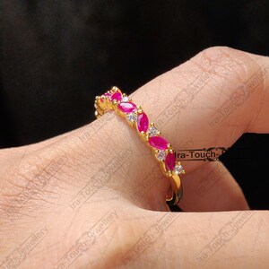 Op de afbeelding: Een gouden bandring met een rij roze ovale edelstenen en kleinere heldere ronde edelstenen. De ring zit aan een vinger.