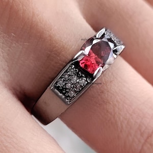May include: A silver ring with a large, oval-cut, deep red gemstone as the centerpiece. The ring features a row of small, clear stones on each side of the main stone, and the band has a dark, textured finish. The ring is shown on a finger.