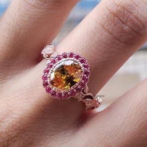 May include: A rose gold ring featuring a large, oval-cut yellow gemstone surrounded by a halo of small, round, red gemstones. The ring's band has intricate floral detailing and small accent stones.