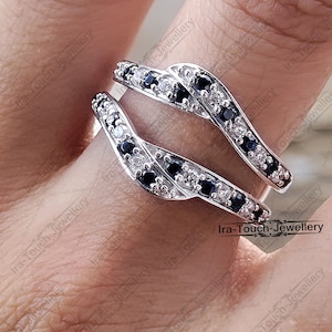 May include: A silver ring with two bands intertwined, set with black and white cubic zirconia stones. The ring is worn on a finger.