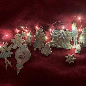 May include: A selection of Christmas decorations, including a fairy, gingerbread man, Santa Claus, house, and star, are displayed on a red surface. The ornaments are lit by a string of small, warm white lights, creating a festive holiday scene.