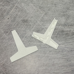 May include: Two white, T-shaped plastic pieces. One piece has a decorative design on the side. The other piece has a circular design on the base. The pieces are on a gray, textured surface.