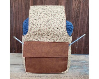 May include: A beige and brown yarn holder with a brown suede pocket and two knitting needles sticking out of the top. The yarn holder is filled with blue yarn.