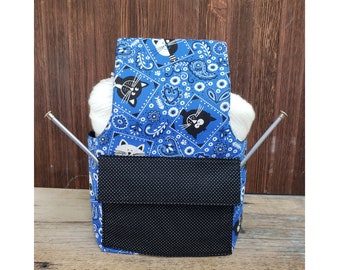 May include: Blue and white patterned yarn holder with two knitting needles sticking out of the top. The holder has a black pocket on the front with a white polka dot pattern.