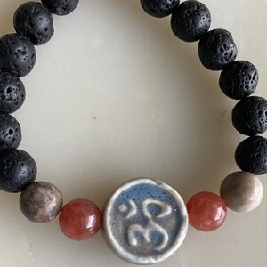 May include: A black lava rock bracelet with a blue and white ceramic charm featuring the Om symbol. The bracelet also includes red and brown beads.