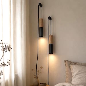 Op de afbeelding: Twee aan de muur gemonteerde hanglampen met cilindrische kappen, bestaande uit een bovenste deel van natuurlijk hout en een onderste deel in matzwart. Elke lamp hangt aan een zwart snoer dat is bevestigd aan een ronde zwarte muurbevestiging.