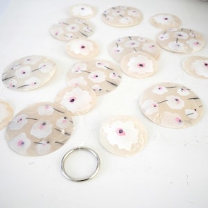 K&ouml;nnte beinhalten: Eine Ansammlung kreisf&ouml;rmiger, durchscheinender Muschelscheiben mit einem aufgemalten wei&szlig;-rosa Blumenmuster und d&uuml;nnen schwarzen Stielen. Die Scheiben sind mit transparentem Faden verbunden und haben unten einen silberfarbenen Metallring.