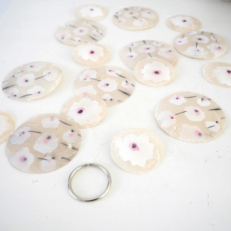 K&ouml;nnte beinhalten: Eine Ansammlung kreisf&ouml;rmiger, durchscheinender Muschelscheiben mit einem aufgemalten wei&szlig;-rosa Blumenmuster und d&uuml;nnen schwarzen Stielen. Die Scheiben sind mit transparentem Faden verbunden und haben unten einen silberfarbenen Metallring.
