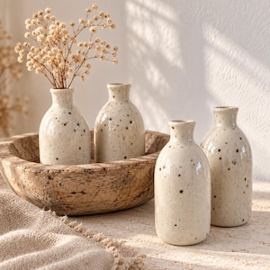 May include: Four small, off-white ceramic bud vases with dark brown speckles. Two vases are placed inside a large, round, weathered wooden bowl, and two are positioned in front. One vase contains a cluster of dried, light-coloured baby's breath flowers.