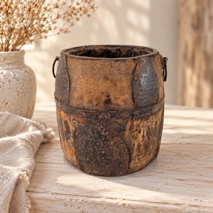 Op de afbeelding: Een cilindrische houten emmer met een verweerde, donkerbruine afwerking, voorzien van verticale metalen banden en donkere, getextureerde metalen platen die met klinknagels zijn bevestigd. De emmer heeft twee kleine metalen ringen aan de zijkanten.
