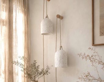 May include: Two bell-shaped pendant lights with a speckled off-white finish, each suspended by a natural fiber rope from a cylindrical wooden wall hook. The lights hang at different heights against a plain wall, featuring a simple, functional design.