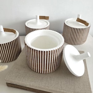 May include: Four handmade ceramic containers with lids. The containers are cylindrical with vertical brown and white stripes. One lid is removed, revealing the interior. Each container has a white lid with a small handle. Small brown tags are attached to the containers.
