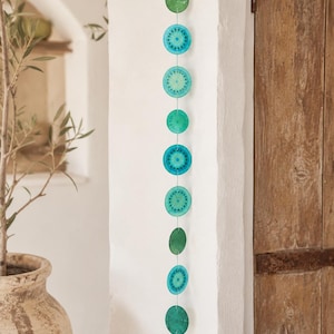 Peut inclure: Une décoration suspendue verticale composée d'une série de onze disques circulaires dans des tons variés de turquoise, de bleu sarcelle et de vert. Les disques sont reliés par un fil fin et présentent des motifs complexes, suspendus à un anneau.