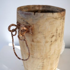 Puede incluir: Un contenedor de madera cilíndrico alto con un asa de metal oxidado. El contenedor está hecho de madera de color claro y tiene un aspecto desgastado y rústico.