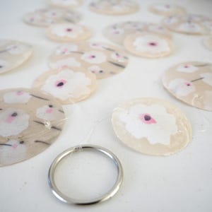 K&ouml;nnte beinhalten: Eine dekorative Girlande mit mehreren durchscheinenden, kreisf&ouml;rmigen Scheiben aus hellbeigem Material. Jede Scheibe ist mit einem sich wiederholenden Muster aus wei&szlig;en Blumen mit kleinen rosa Zentren bedruckt. Die Scheiben sind durch d&uuml;nnen, klaren Nylonfaden verbunden.