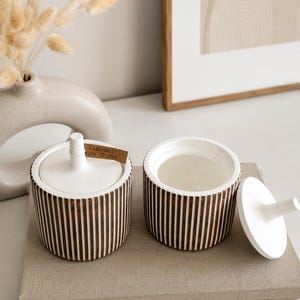 Puede incluir: Dos tarros cilíndricos de cerámica con exteriores de color marrón oscuro con estrías verticales e interiores blancos lisos. Cada tarro tiene una tapa blanca a juego con un asa central. Una pequeña etiqueta marrón dice HANDMADE WITH LOVE.