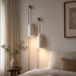 May include: Two cylindrical pendant lights with textured off-white shades suspended from wooden wall hooks by tan fabric cords. The lights emit a warm glow downwards, creating a soft illumination against the wall in a bedroom setting.