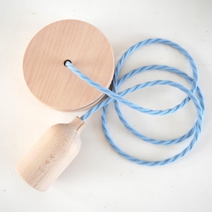 Puede incluir: Una lámpara colgante de madera clara con una base de madera a juego y un cable de tela trenzada azul claro. El cable está en bucle y la lámpara está sobre un fondo blanco.