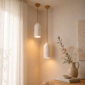May include: Two cylindrical pendant lights with textured white shades and light brown cords suspended from the ceiling. The lights emit a warm glow, illuminating the space below where a ceramic pitcher and stacked books are placed on a surface.
