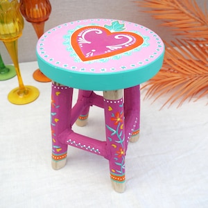 Puede incluir: Un pequeño taburete de madera pintado a mano con una parte superior redonda rosa con un diseño de corazón. El borde es turquesa y las patas son magenta con detalles florales. Es una pieza decorativa.